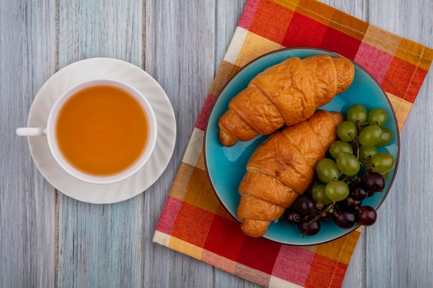 Kostenloses Foto draufsicht von croissants mit trauben im teller auf kariertem stoff und tasse heißem wirbel auf hölzernem hintergrund