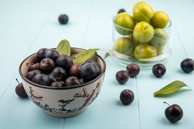 Draufsicht von dunkelvioletten sauren Schlehen auf einer Schüssel mit frischen grünen Kirschpflaumen auf einem Glasglas auf einem blauen Hintergrund