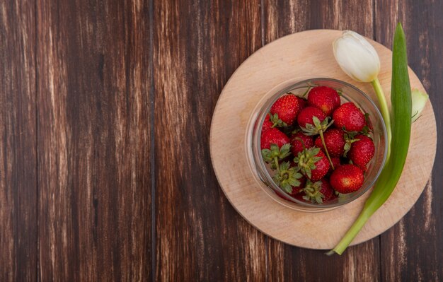 Draufsicht von Erdbeeren in Schüssel und Blume auf Schneidebrett auf hölzernem Hintergrund mit Kopienraum