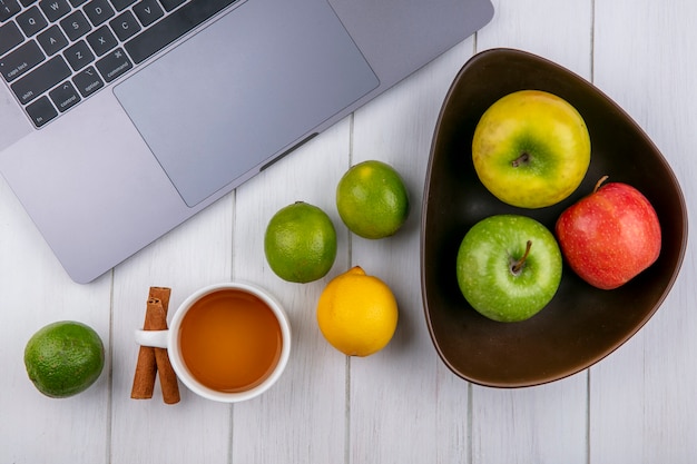 Kostenloses Foto draufsicht von farbigen äpfeln in einer schüssel mit laptop limetten eine tasse tee und zimt auf einer weißen oberfläche
