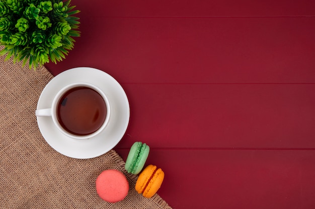 Draufsicht von farbigen Macarons mit einer Tasse Tee auf einer beigen Serviette auf einer roten Oberfläche
