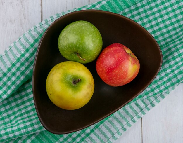 Draufsicht von farbigen Äpfeln in einer Schüssel auf einem grünen karierten Handtuch auf einer weißen Oberfläche