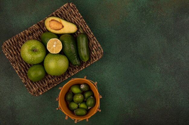 Draufsicht von Feijoas auf einem Eimer mit Avocados-Gurke der grünen Äpfel auf einem Weidentablett auf einer grünen Oberfläche mit Kopierraum
