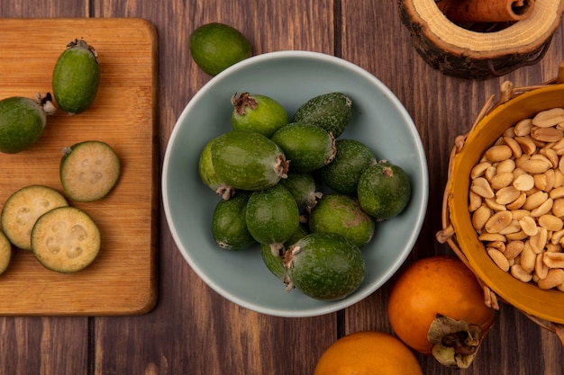 Draufsicht von Feijoas auf einer Schüssel mit Scheiben von Feijoas auf einem hölzernen Küchenbrett mit Erdnüssen auf einem Eimer auf einem hölzernen Hintergrund