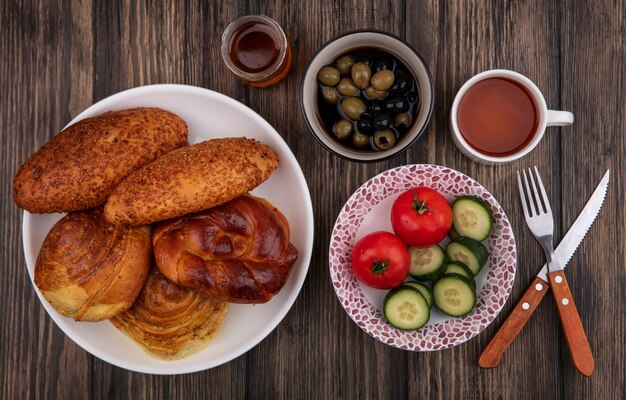 Draufsicht von frischen Brötchen auf einem weißen Teller mit Gemüse mit Oliven auf einer Schüssel auf einem hölzernen Hintergrund