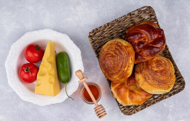 Draufsicht von frischen Brötchen auf Weidenschale mit Gemüse und Käse auf einem weißen Teller mit Honig auf weißem Hintergrund