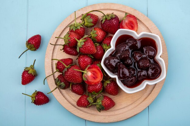 Draufsicht von frischen Erdbeeren mit einer Erdbeermarmelade auf einer Schüssel auf einem hölzernen Küchenbrett auf einem blauen Hintergrund