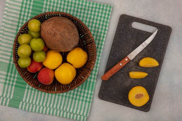 Draufsicht von frischen Früchten wie grüner Kirschpflaumenpfirsich-Kokosnuss auf einem Eimer mit Pfirsichscheiben auf einem Küchenschneidebrett mit Messer auf weißem Hintergrund