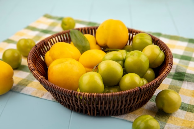 Draufsicht von frischen gelben Pfirsichen auf einem Eimer mit grünen Kirschpflaumen auf einem karierten Stoff auf einem blauen Hintergrund