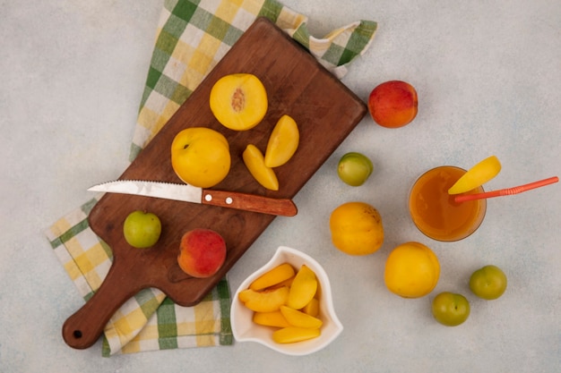 Draufsicht von frischen gelben Pfirsichen auf einem hölzernen Küchenbrett auf einer karierten Tischdecke mit Messer mit frischem Pfirsichsaft auf einem Glas auf einem weißen Hintergrund