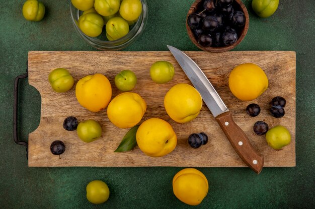 Draufsicht von frischen gelben Pfirsichen auf einem hölzernen Küchenbrett mit grünen Kirschpflaumen mit Messer mit Schlehen auf einer Holzschale auf einem grünen Hintergrund