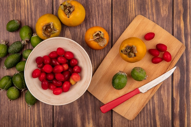 Draufsicht von frischen Kornelkirschen auf einer Schüssel mit Kaki und Feijoas auf einem hölzernen Küchenbrett mit Messer auf einer hölzernen Oberfläche