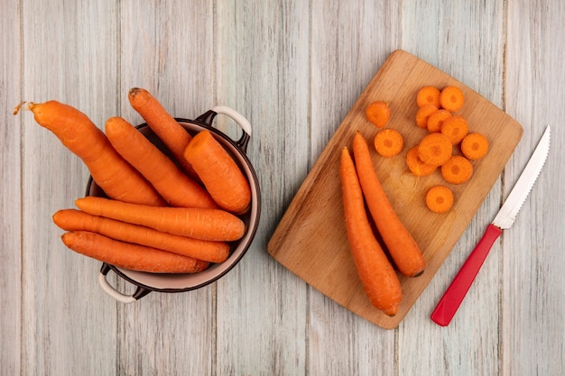 Kostenloses Foto draufsicht von frischen orange karotten auf einem hölzernen küchenbrett mit messer mit karotten auf einer schüssel auf einer grauen holzoberfläche