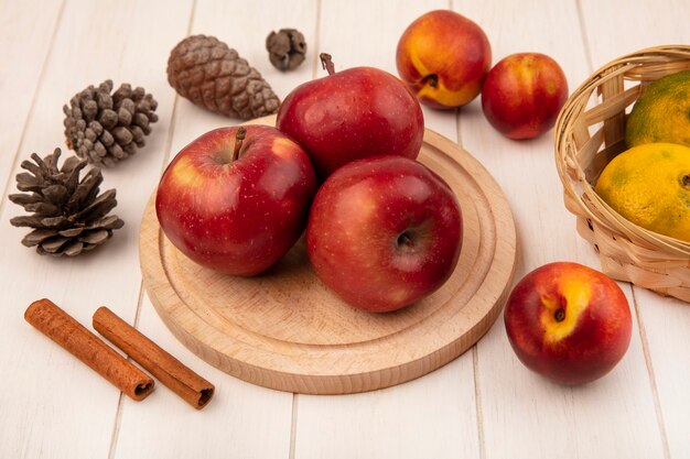 Draufsicht von frischen Äpfeln auf einem hölzernen Küchenbrett mit Mandarinen auf einem Eimer mit Pfirsichen Zimtstangen und Tannenzapfen lokalisiert auf einer weißen Holzwand