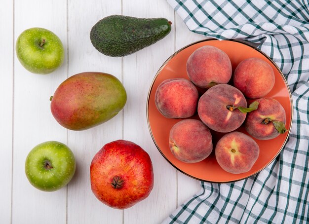 Draufsicht von frischen Pfirsichen auf Schüssel auf karierter Tischdecke mit Apfel-Mango-Granatapfel lokalisiert auf Weiß