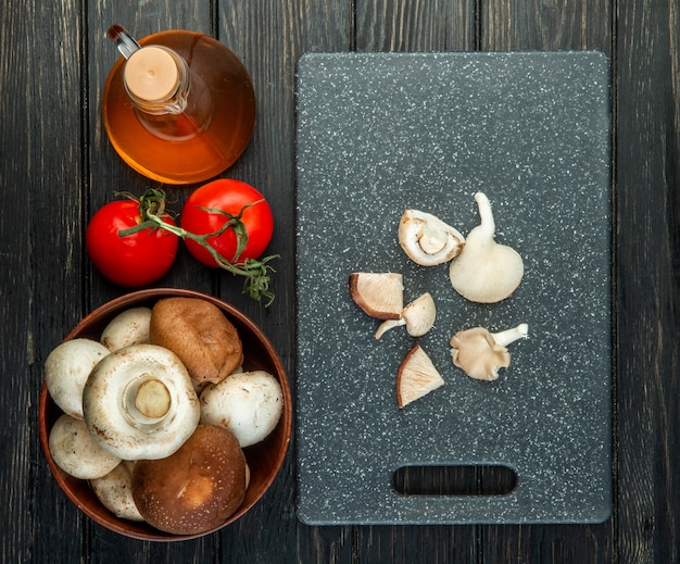 Draufsicht von frischen Pilzen in einer Holzschüsselflasche des Olivenöls und von frischen Tomaten geschnittenen Pilzen auf schwarzem Schneidebrett auf Schwarz