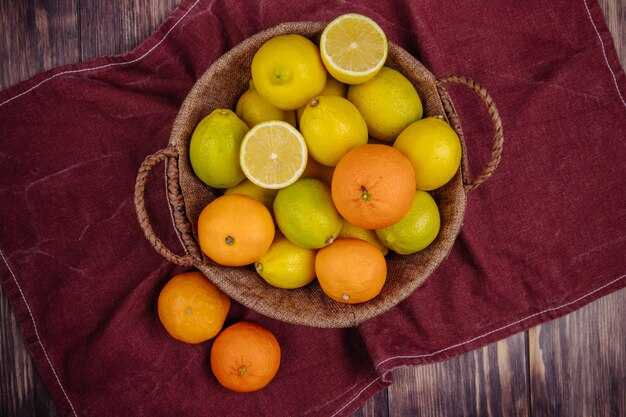 Draufsicht von frischen reifen Zitronen und Orangen in einem Weidenkorb auf dunkelrotem Stoff auf rustikalem