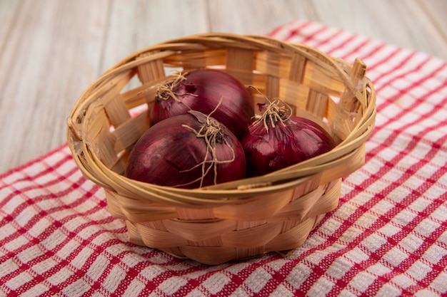 Draufsicht von frischen roten Zwiebeln auf einem Eimer auf einem rot karierten Tuch auf einer grauen Holzoberfläche