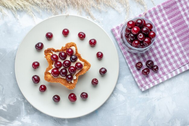 Draufsicht von frischen Sauerkirschen innerhalb Platte mit sternförmigem cremigem Kuchen auf hellweißem Schreibtisch, Fruchtsauer-Sommerkuchen-Keks