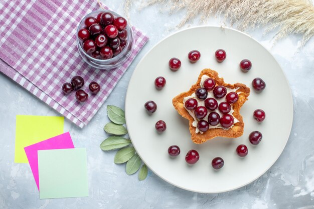 Draufsicht von frischen Sauerkirschen innerhalb Platte mit sternförmigem cremigem Kuchen auf weißem weißem Schreibtisch, Obstsauer-Sommerkuchen-Keks