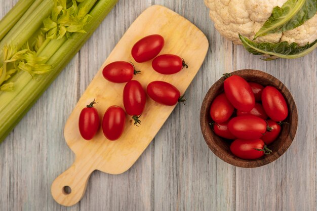 Draufsicht von frischen Tomaten auf einem hölzernen Küchenbrett mit Tomaten auf einer hölzernen Schüssel mit Blumenkohl und Sellerie lokalisiert auf einer grauen Holzoberfläche