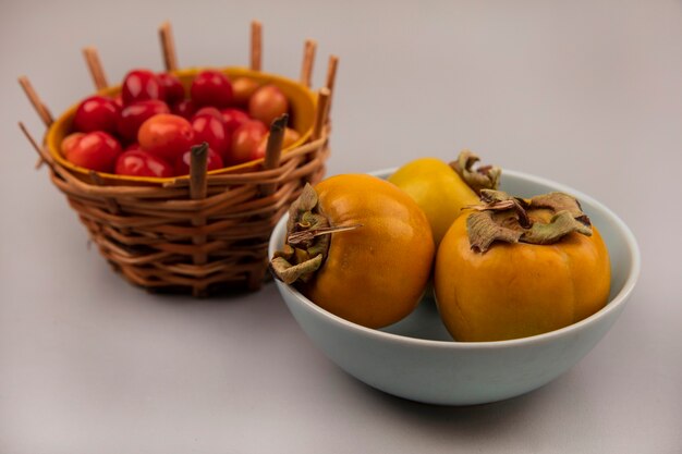 Draufsicht von frischen und gesunden Kornelkirschenfrüchten auf einem Eimer mit Kakifruchtfrüchten auf einer Schüssel auf einem grauen Hintergrund