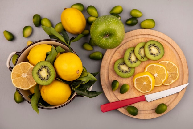 Draufsicht von frischen Zitronen auf einer Schüssel mit Kiwi- und Zitronenscheiben auf einem hölzernen Küchenbrett mit Messer mit Kinkans und Apfel lokalisiert auf einer weißen Wand