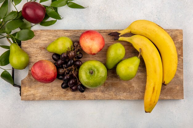 Draufsicht von Früchten als Birnenapfel-Traubenpfirsichbanane auf Schneidebrett mit Blättern auf weißem Hintergrund