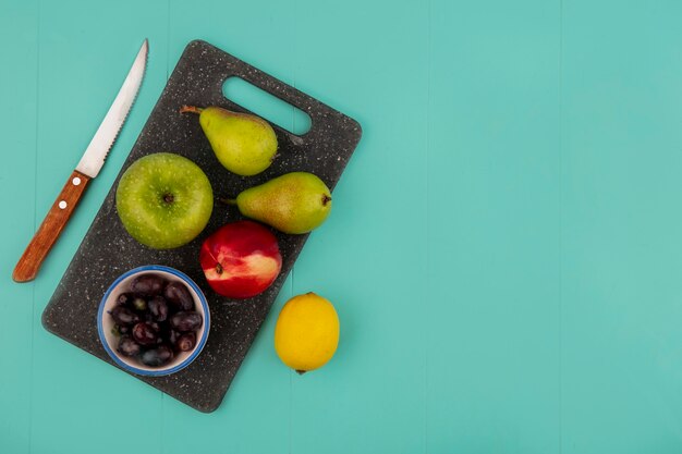 Draufsicht von Früchten als Birnenpfirsichapfel und Traubenbeeren auf Schneidebrett mit Zitrone und Messer auf blauem Hintergrund mit Kopienraum