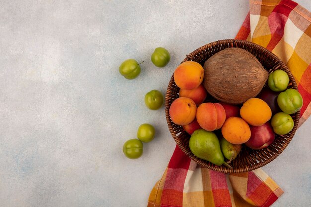 Draufsicht von Früchten als Kokosnuss-Aprikosen-Pfirsichbirne im Korb auf kariertem Stoff mit Pflaumen auf weißem Hintergrund mit Kopienraum