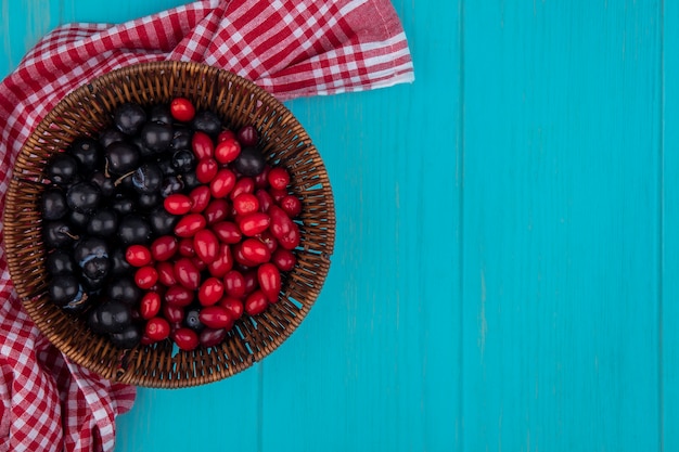 Draufsicht von Früchten als Kornelkirsche und Schlehenbeeren im Korb auf kariertem Stoff auf blauem Hintergrund mit Kopienraum