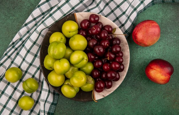 Draufsicht von Früchten als Pflaumen und Kirschen in Platte auf kariertem Stoff mit Pfirsichen auf grünem Hintergrund