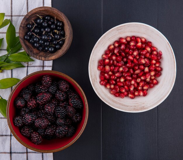 Draufsicht von Früchten als Schalen von Sloe und Brombeere mit Blättern auf kariertem Stoff und Schüssel Granatapfelbeeren auf schwarzer Oberfläche