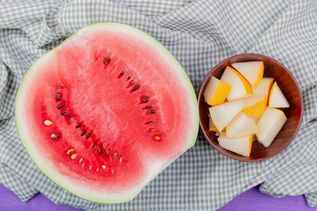 Draufsicht von Früchten als Wassermelonenhälfte und Schüssel geschnittene Melone auf kariertem Stoff und lila Hintergrund