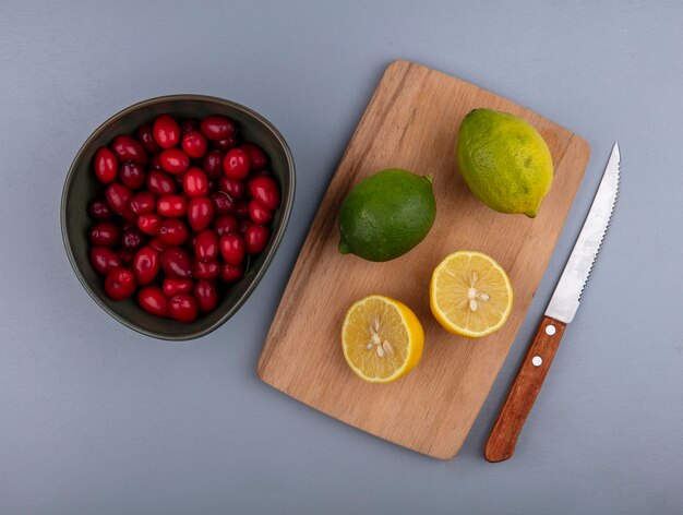 Draufsicht von ganzen und halb geschnittenen Zitronen auf Schneidebrett und Schüssel von Kornelkirschenbeeren mit Messer auf grauem Hintergrund