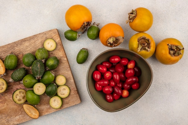 Draufsicht von ganzen und halbierten reifen Feijoas lokalisiert auf einem hölzernen Küchenbrett mit Kakis und Kornelkirschen auf einer Schüssel auf einer grauen Oberfläche