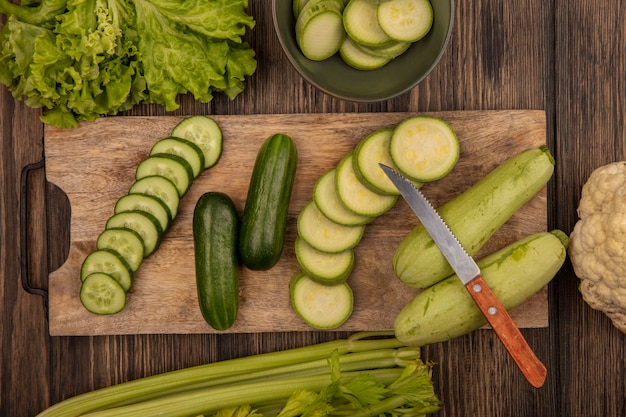 Draufsicht von gehackten Gurken und Zucchini lokalisiert auf einem hölzernen Küchenbrett mit Messer mit Salatsellerie und Blumenkohl lokalisiert auf einer Holzwand