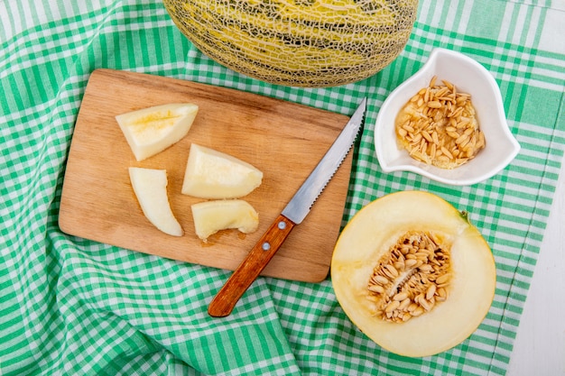 Draufsicht von gehackten Melonenscheiben auf hölzernem Küchenbrett mit Messer mit Samen auf weißer Schüssel auf gelber karierter Tischdecke