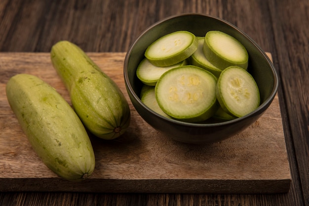 Draufsicht von gehackten Zucchini auf einer Schüssel mit ganzen Zucchini lokalisiert auf einem hölzernen Küchenbrett auf einer Holzwand