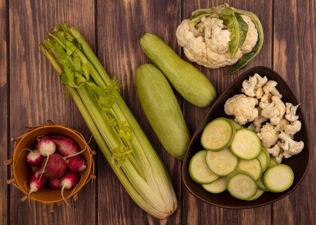 Draufsicht von gehackten Zucchini und Blumenkohlknospen auf einer Schüssel mit Radieschen auf einem Eimer mit ganzem Zucchinisellerie und Blumenkohl isoliert auf einer Holzoberfläche