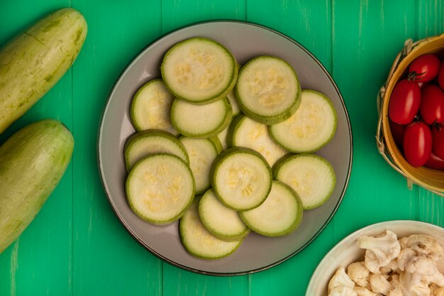 Draufsicht von gehackten Zucchinischeiben auf einem Teller mit Pflaumentomaten auf einem Eimer mit Blumenkohlblüten auf einer Schüssel mit Zucchini lokalisiert auf einer grünen Holzwand