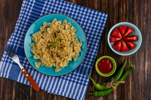 Draufsicht von gekochten Nudeln auf einem blauen Teller auf einem blau karierten Handtuch mit einem Gabel-Tomaten-Ketchup und Chilischoten auf einer Holzoberfläche