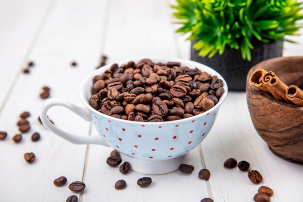 Draufsicht von gerösteten Kaffeebohnen auf einer Tasse mit Zimtstangen auf einer Holzschale auf einem weißen hölzernen Hintergrund