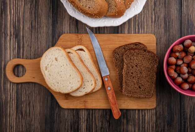 Draufsicht von geschnittenem Brot als gesätem braunem Cob Roggen und weißen in Platte auf kariertem Stoff und auf Schneidebrett mit Messer und Schüssel Nüssen auf hölzernem Hintergrund