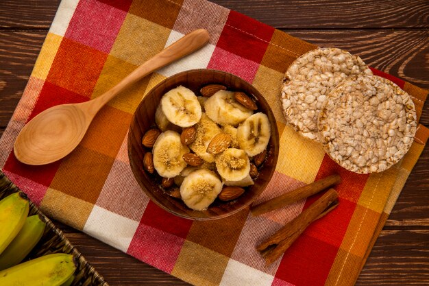 Draufsicht von geschnittenen Bananen mit Mandel in einer Schüssel und Holzlöffeln mit Reiscrackern und Zimtstangen auf rustikalem