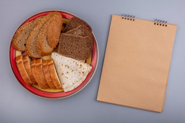 Draufsicht von geschnittenen Broten als gesätes Fladenbrot des braunen Kolbens knusprig und Roggen in Platte mit Notizblock auf grauem Hintergrund mit Kopienraum