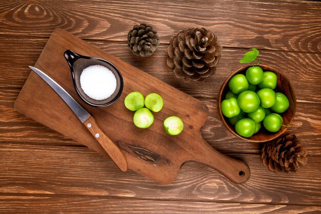 Draufsicht von geschnittenen grünen Pflaumen mit Salz und Küchenmesser auf einem hölzernen Schneidebrett, Zapfen und Pflaumen in einer Schüssel auf rustikalem Holztisch