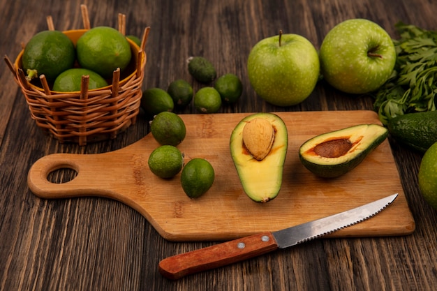 Draufsicht von gesunden Avocados auf einem hölzernen Küchenbrett mit Messer mit Limetten auf einem Eimer mit Äpfeln feijoas und Petersilie lokalisiert auf einem hölzernen Hintergrund
