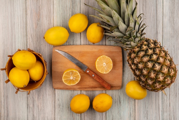 Draufsicht von gesunden Zitronen auf einem Eimer mit halben Zitronen auf einem hölzernen Küchenbrett mit Messer mit Zitronen und Ananas lokalisiert auf einem grauen hölzernen Hintergrund