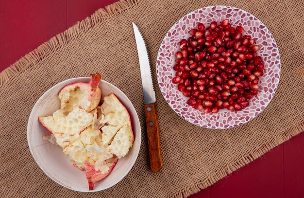 Draufsicht von Granatapfelbeeren in Schüssel und Schüssel Granatapfelschale mit Messer auf Sackleinen auf Bordo-Oberfläche